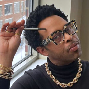 Aaron Brown, wearing glasses matching his gold necklace and bracelets, leaning into his hand hodling a pencil and looking thoughtful.